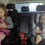 une petite fille et un bébé assis dans une voiture chacun dans leur siège auto avec une petite peluche assis entre eux