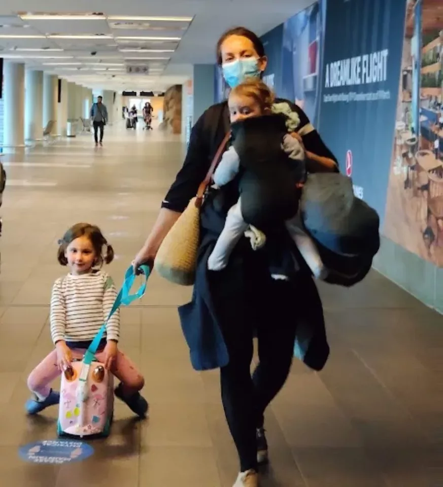 Maman dans un aéroport avec un bébé en porte-bébé et une petite fille sur une valise à roulettes pour enfant