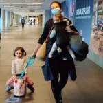 Mother is an airport with a baby in a baby carrier and a little girl on a kids rolling suitcase